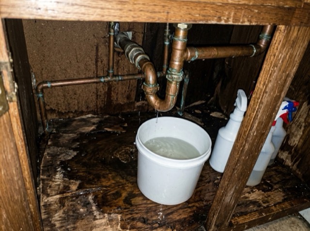 Photo the tenant sent: water dripping from a copper pipe under a kitchen sink into a bucket